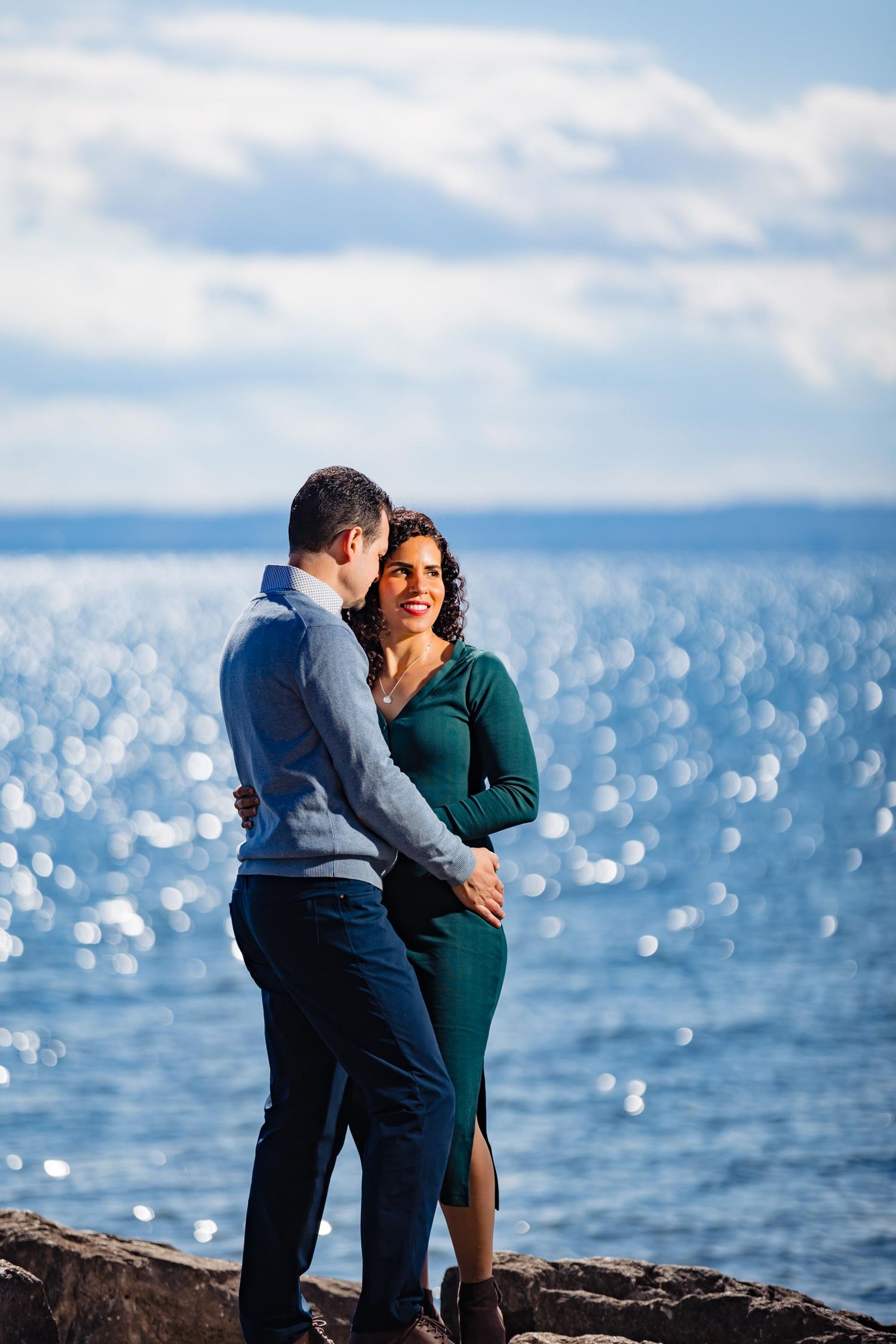 A couple sharing an intimate moment on rocky shores with sparkling water in the background.