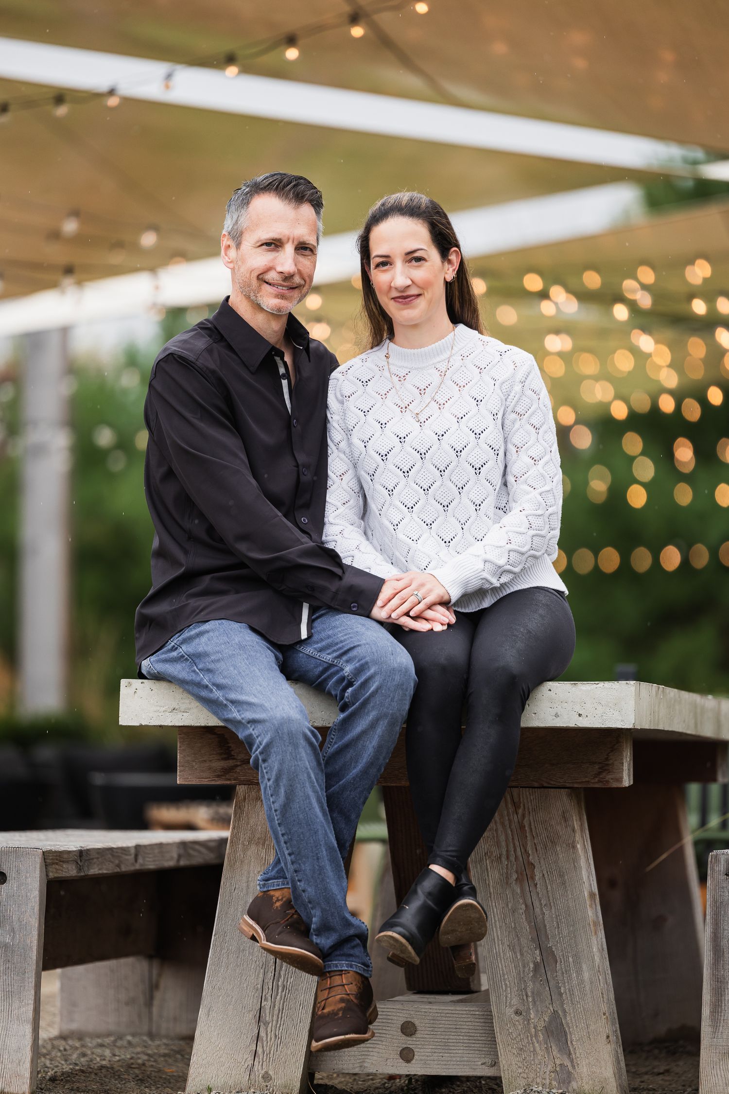 A couple sits together on a wooden bench under string lights during an outdoor engagement photo session.