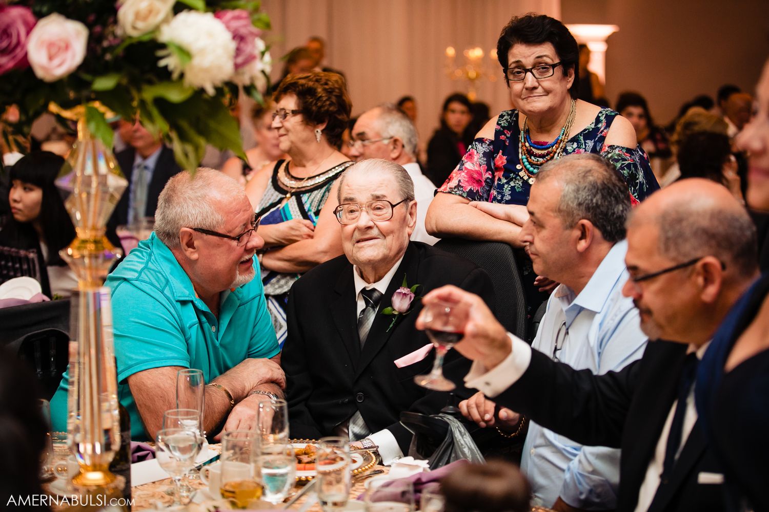 Wedding reception guests seated at tables in an elegant banquet hall with warm lighting and floral centerpieces.