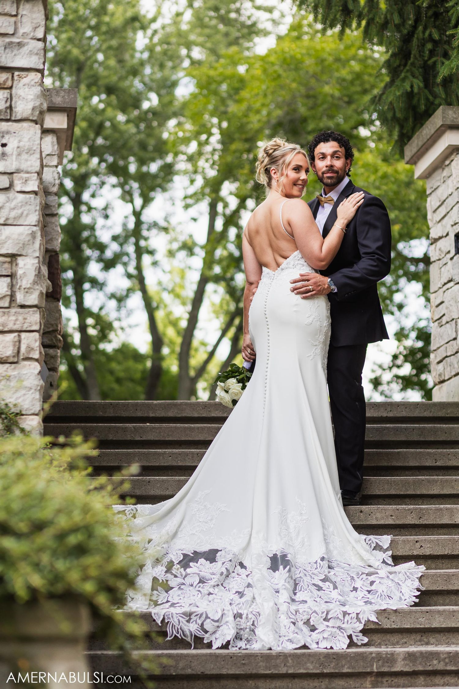 Bride in fitted lace gown with dramatic train poses with partner on stone steps surrounded by greenery.