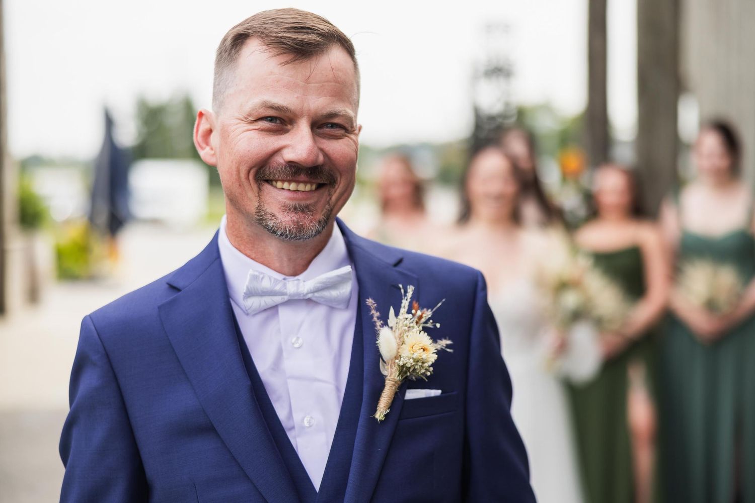 Groom in navy suit with bowtie smiles brightly during outdoor wedding.