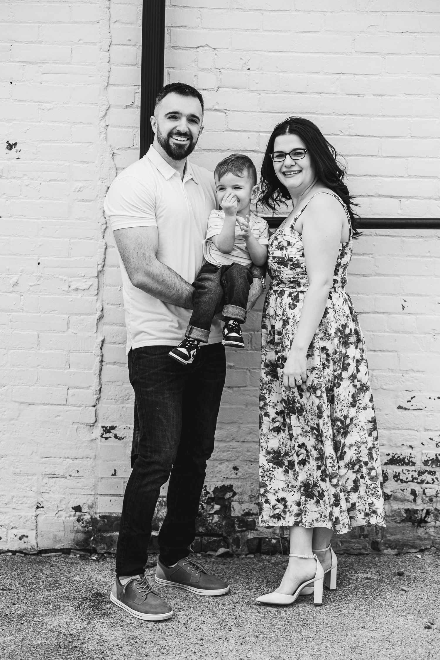 Family of three poses casually against white brick wall in black and white portrait.