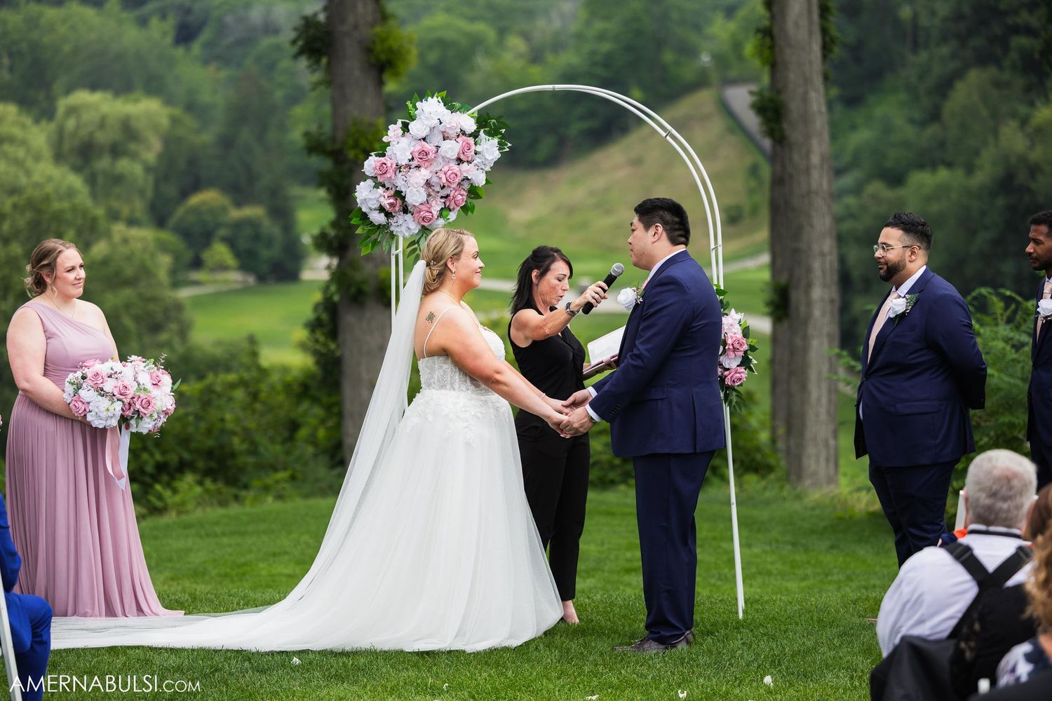 Wedding ceremony under a curved arch decorated with pink floral arrangements in an outdoor garden setting.