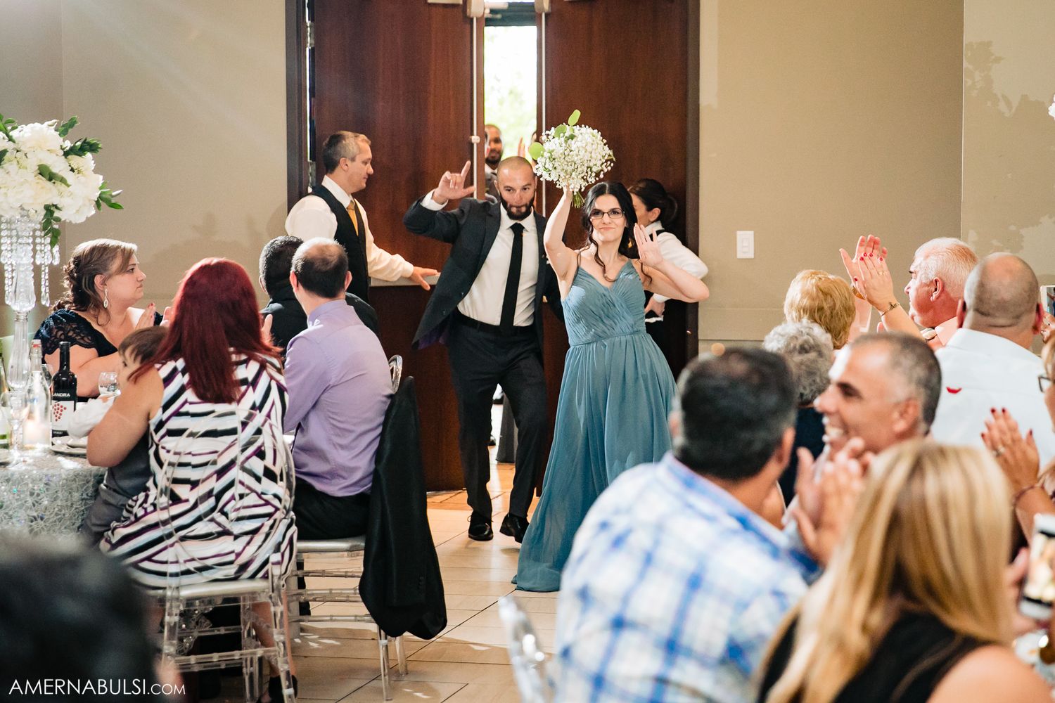 Wedding reception guests celebrate and dance in a banquet hall decorated with white flowers.