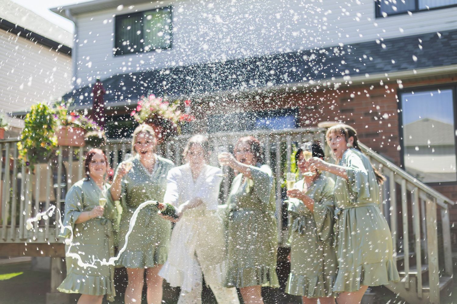Bridesmaids in sage robes spray champagne with bride outdoors in bright sunlight.