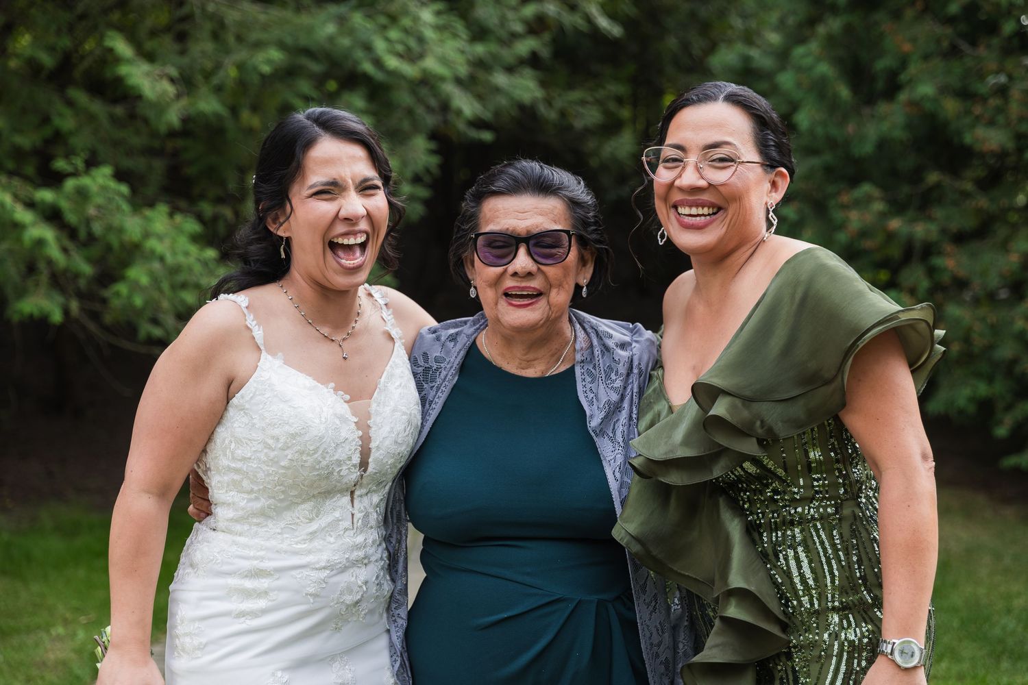 Bride poses with two smiling women in elegant attire at outdoor wedding celebration.
