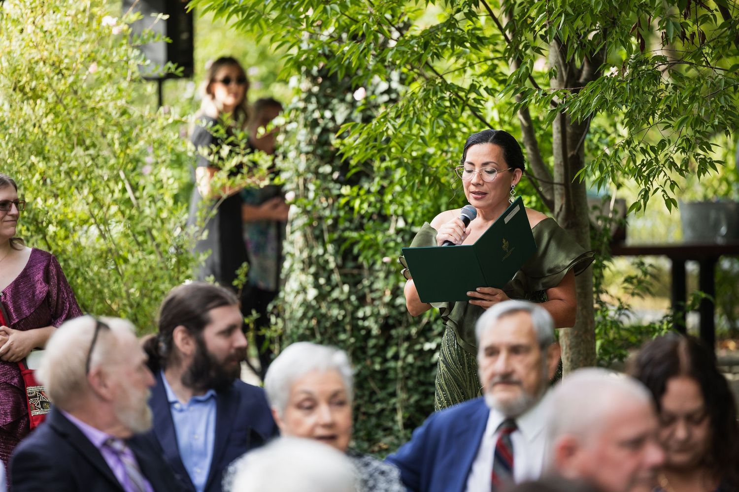 Woman reading from a book while giving a speech at an outdoor wedding ceremony surrounded by greenery.