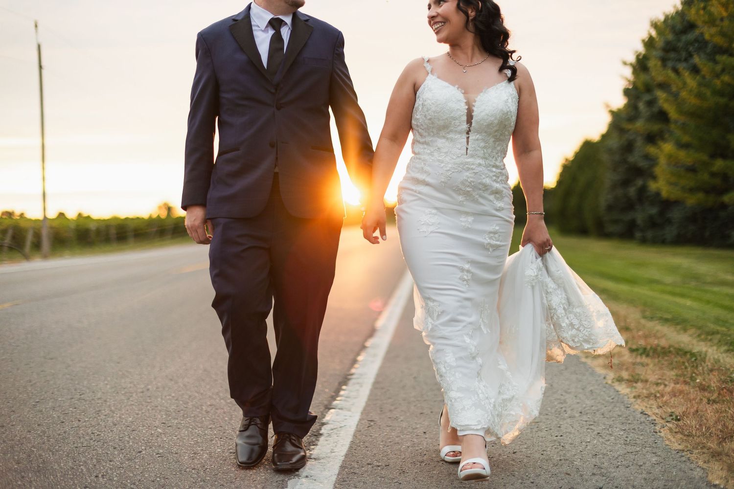 Bride and groom walking hand in hand down a sunlit road at sunset, bride in lace gown, groom in navy suit.