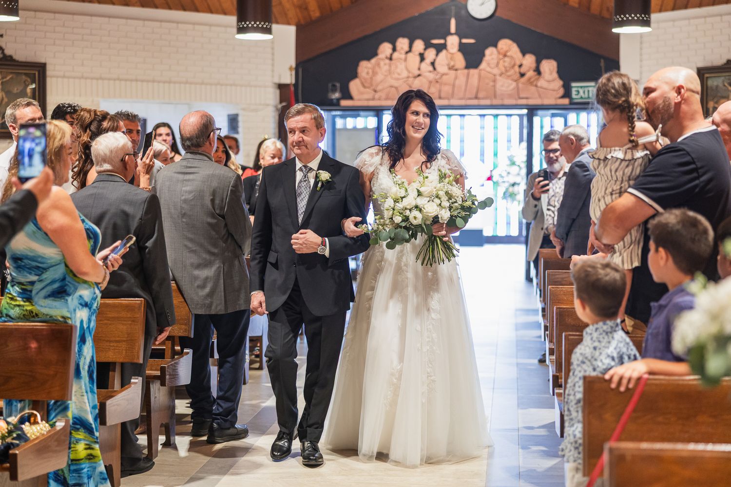 Father escorts bride holding white bouquet down the church aisle on her wedding day.