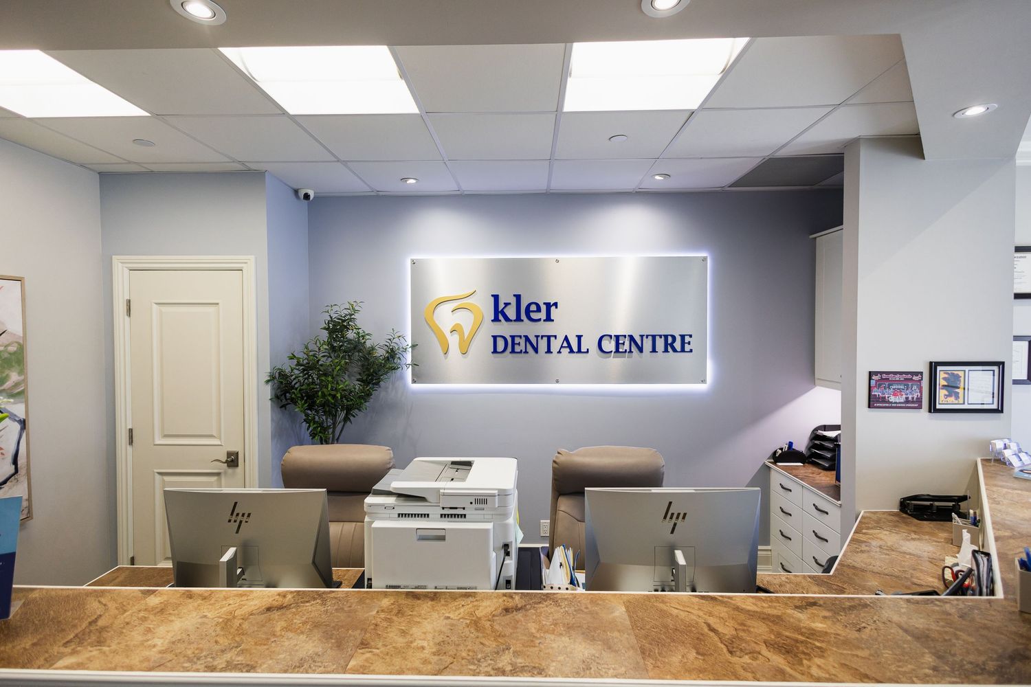 Modern dental office reception area featuring Kler Dental Centre signage, front desk computers, and a welcoming blue interior.