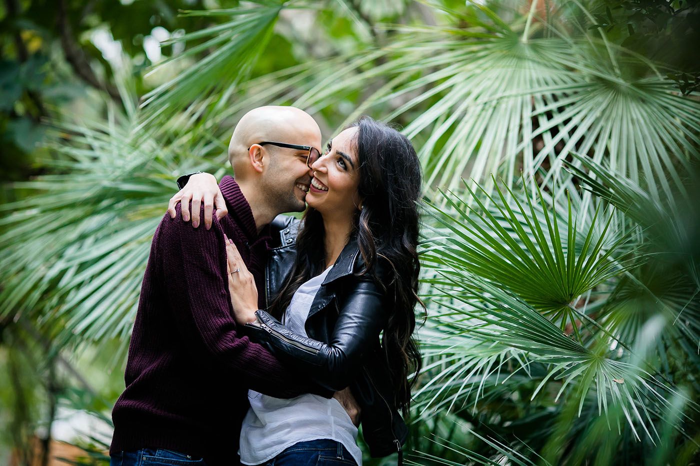 Couple shares tender moment surrounded by palm fronds and tropical plants.