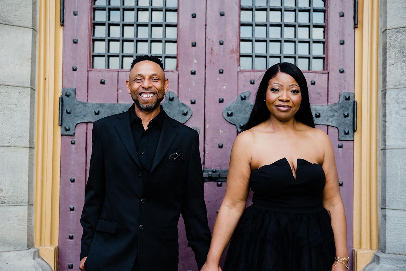 Elegant couple in formal black attire posing in front of stone building with ornate doors.