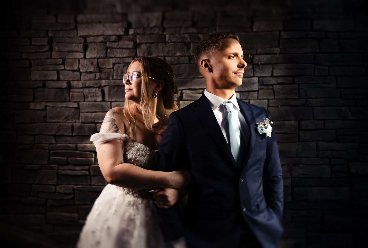 A dramatic portrait of newlyweds against a dark brick wall with moody lighting.