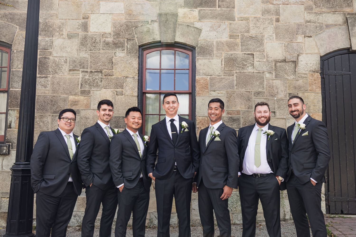 Seven groomsmen in matching dark suits pose together against a stone building wall.