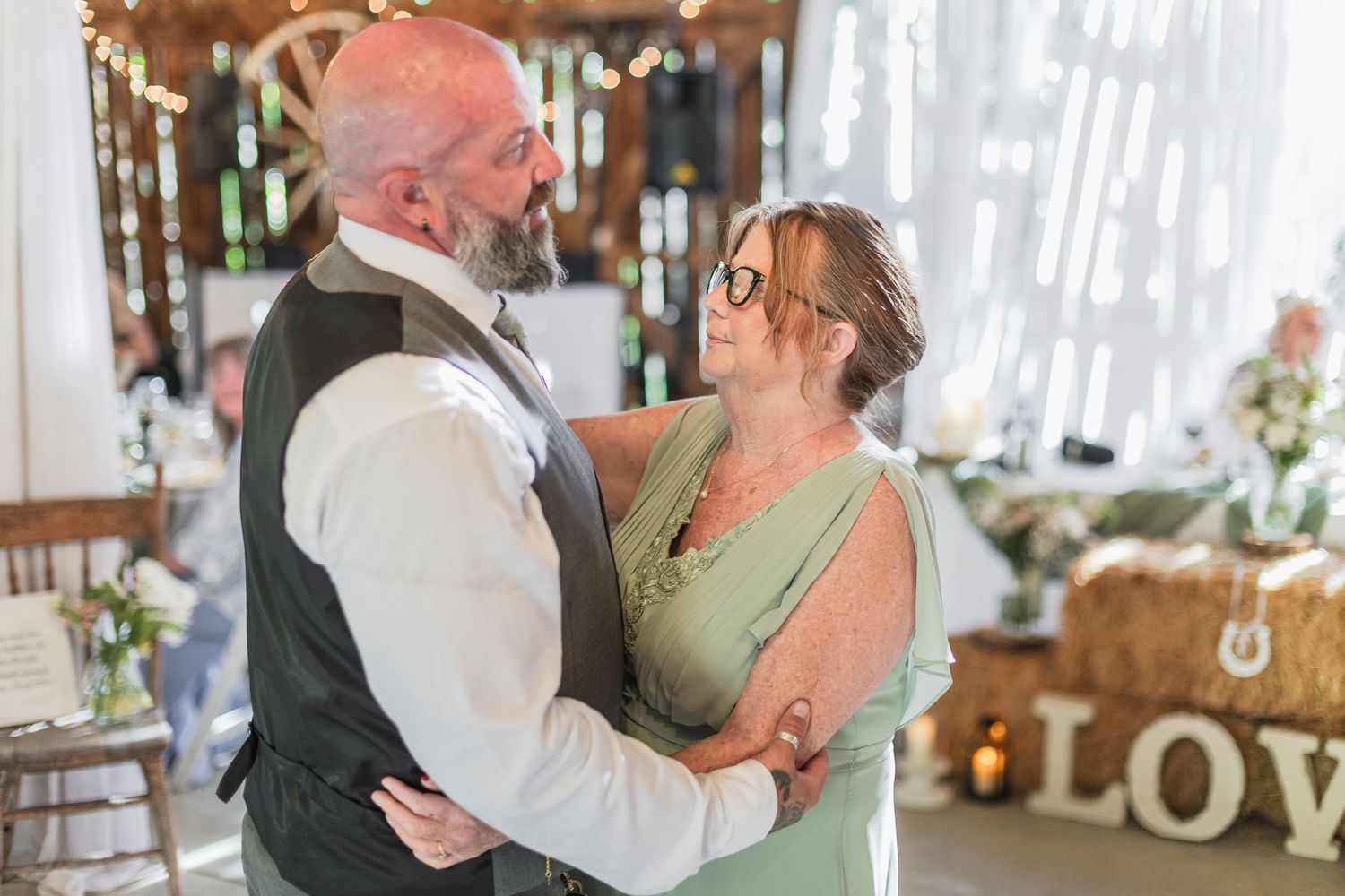 Couple shares romantic dance at rustic barn wedding reception with LOVE sign and hay bales in background.