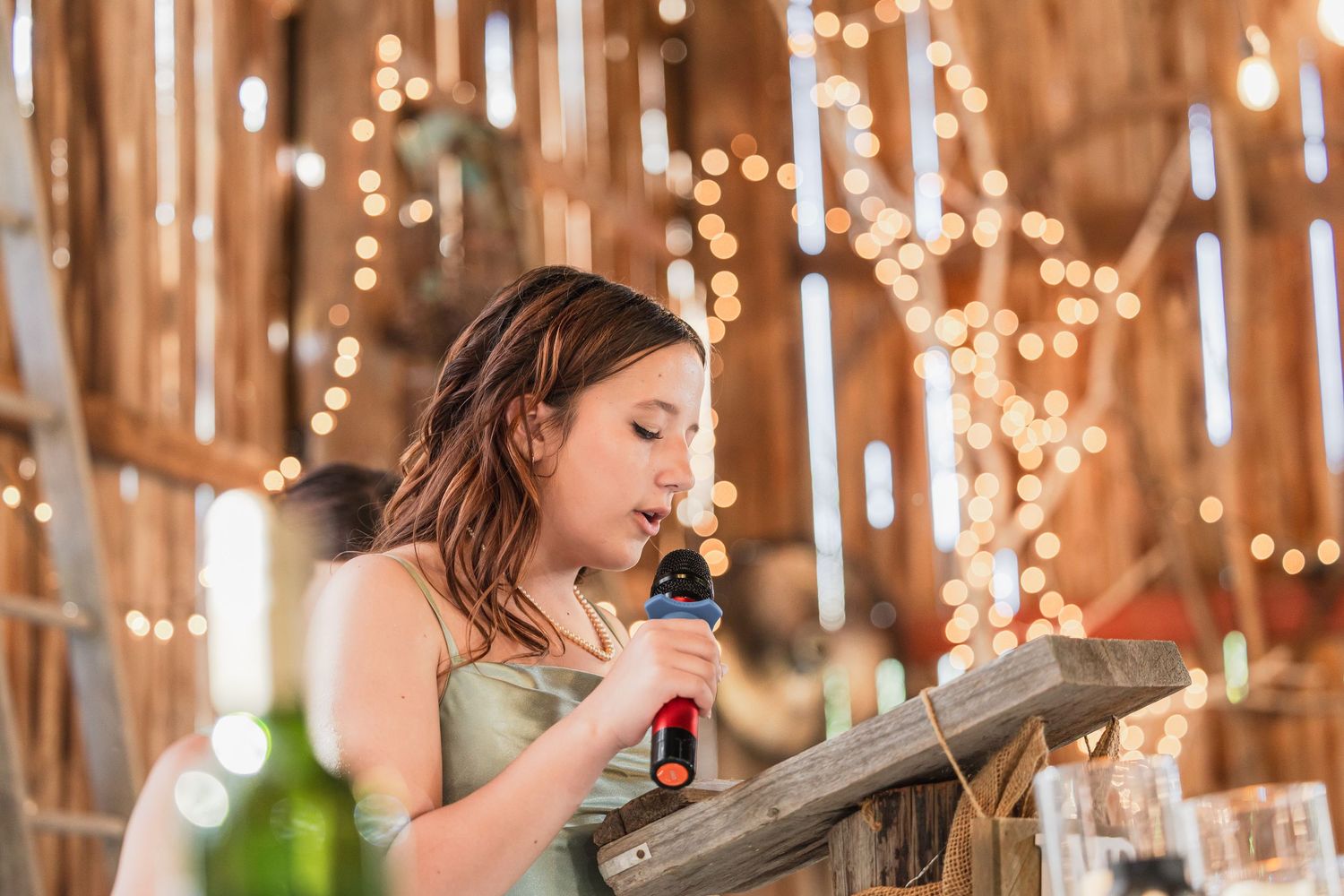 Young girl speaking into a microphone at a rustic barn wedding reception with warm string lights in background.