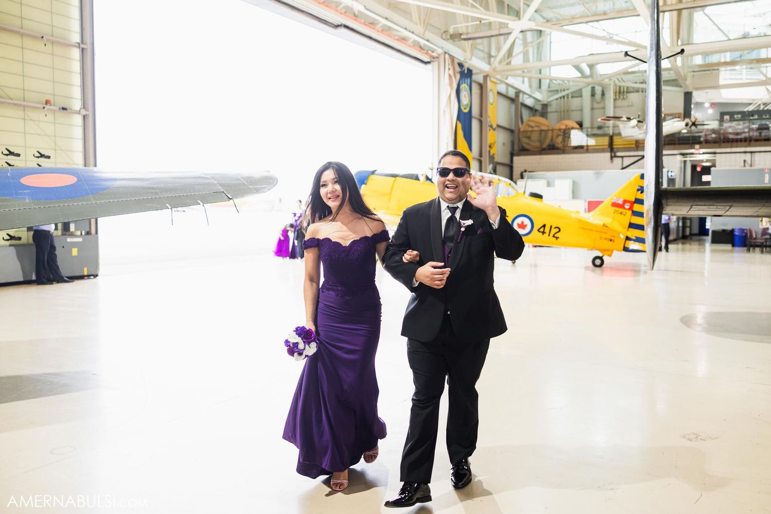 A couple walks through an aviation hangar with a yellow vintage aircraft in the background.
