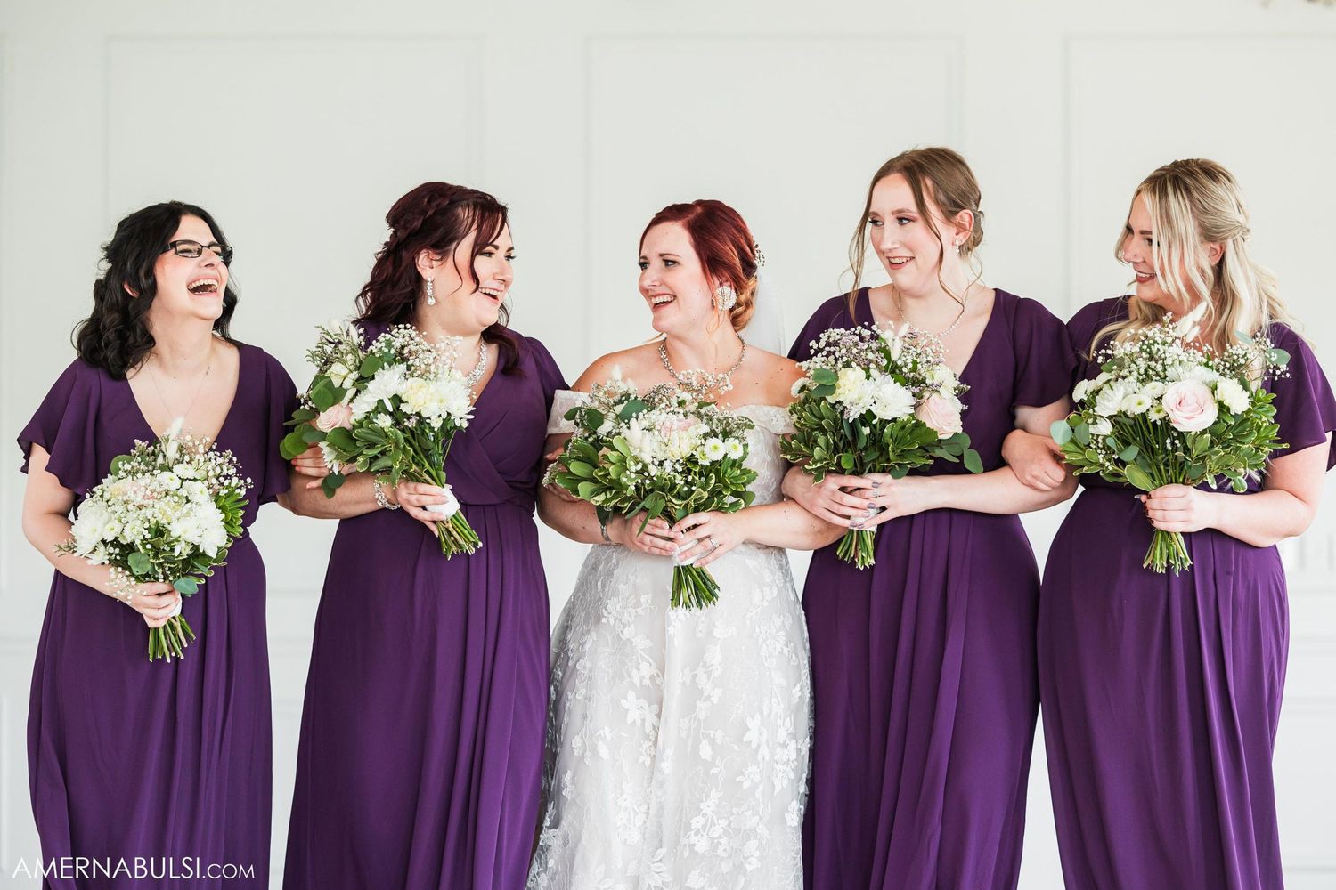 Bridal party in purple dresses holding white bouquets against white wall.