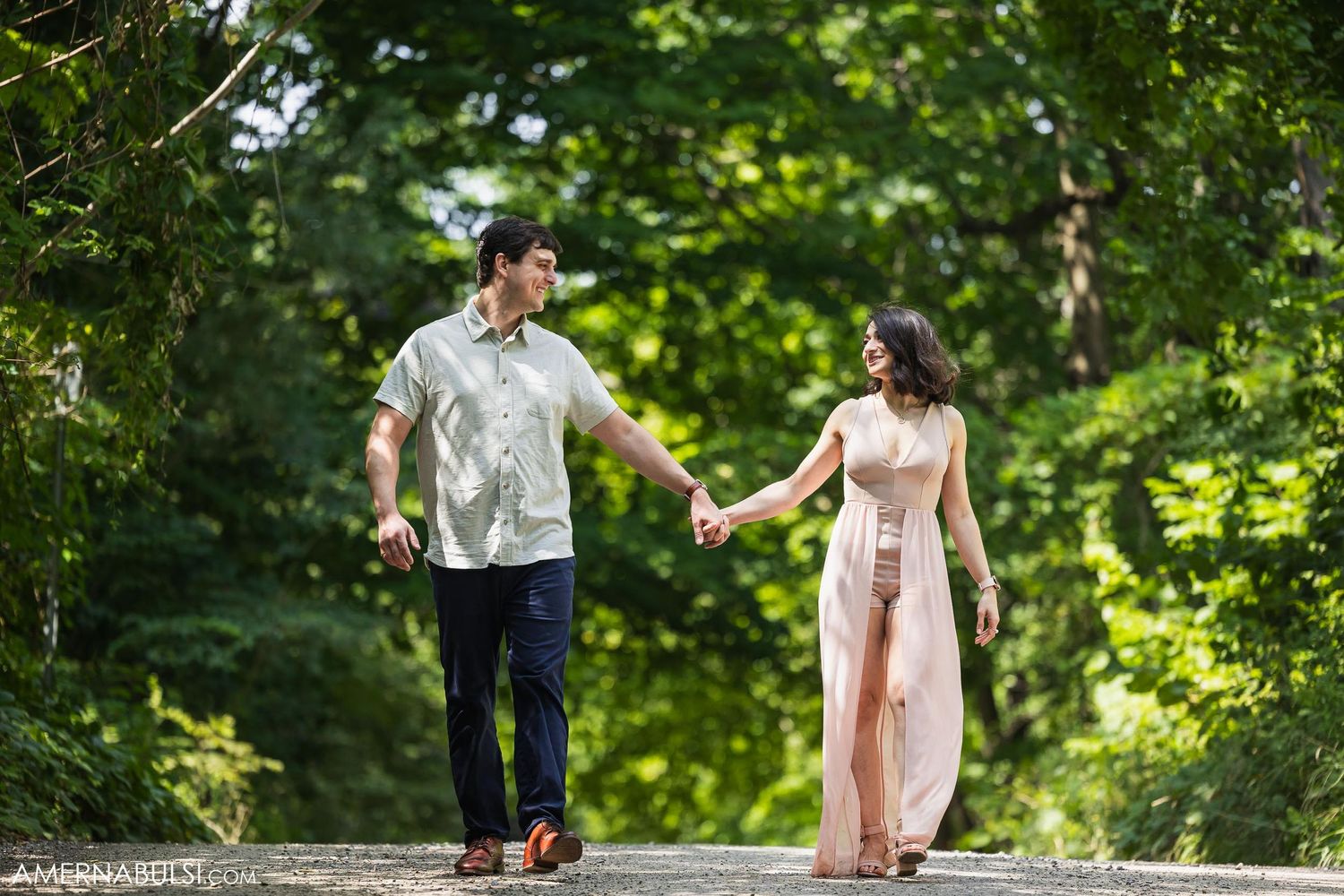 Couple holding hands while walking along sunlit forest path in casual attire.