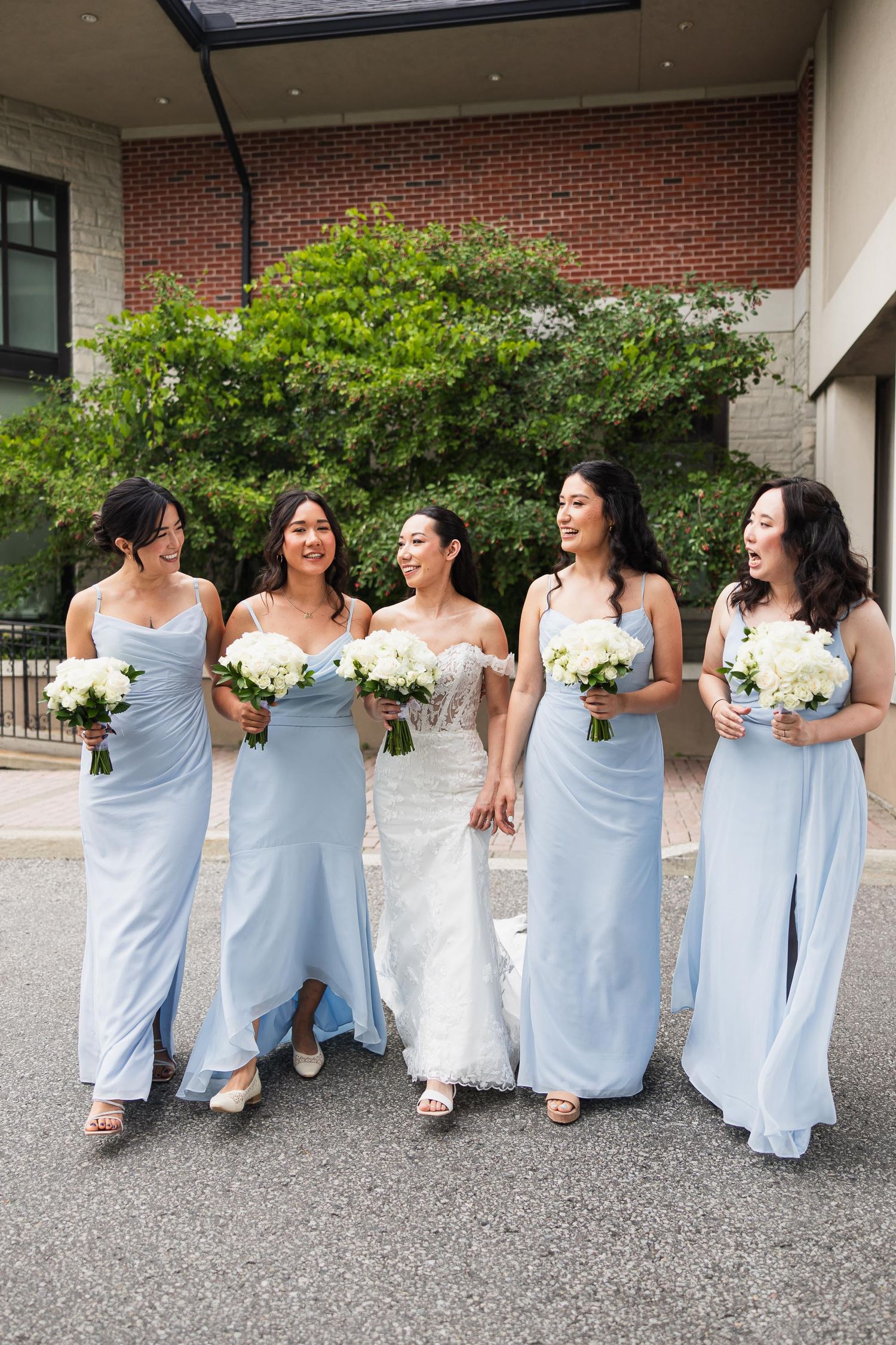 Bridesmaids in pale blue gowns laugh together while holding white floral bouquets.