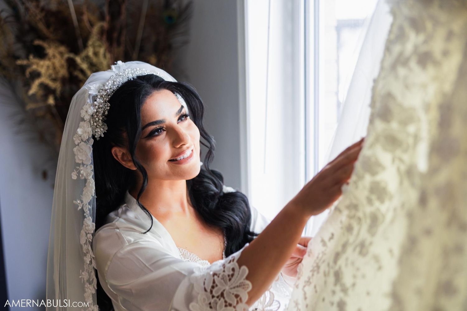 A bride in a white robe and delicate lace veil gazes contemplatively out a bright window.