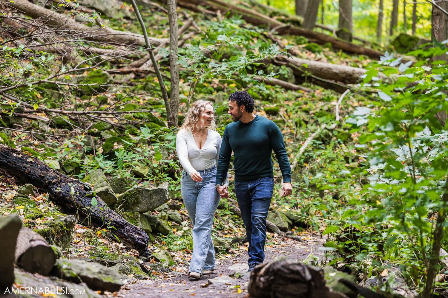 A couple walks hand in hand along a wooded trail surrounded by lush greenery.