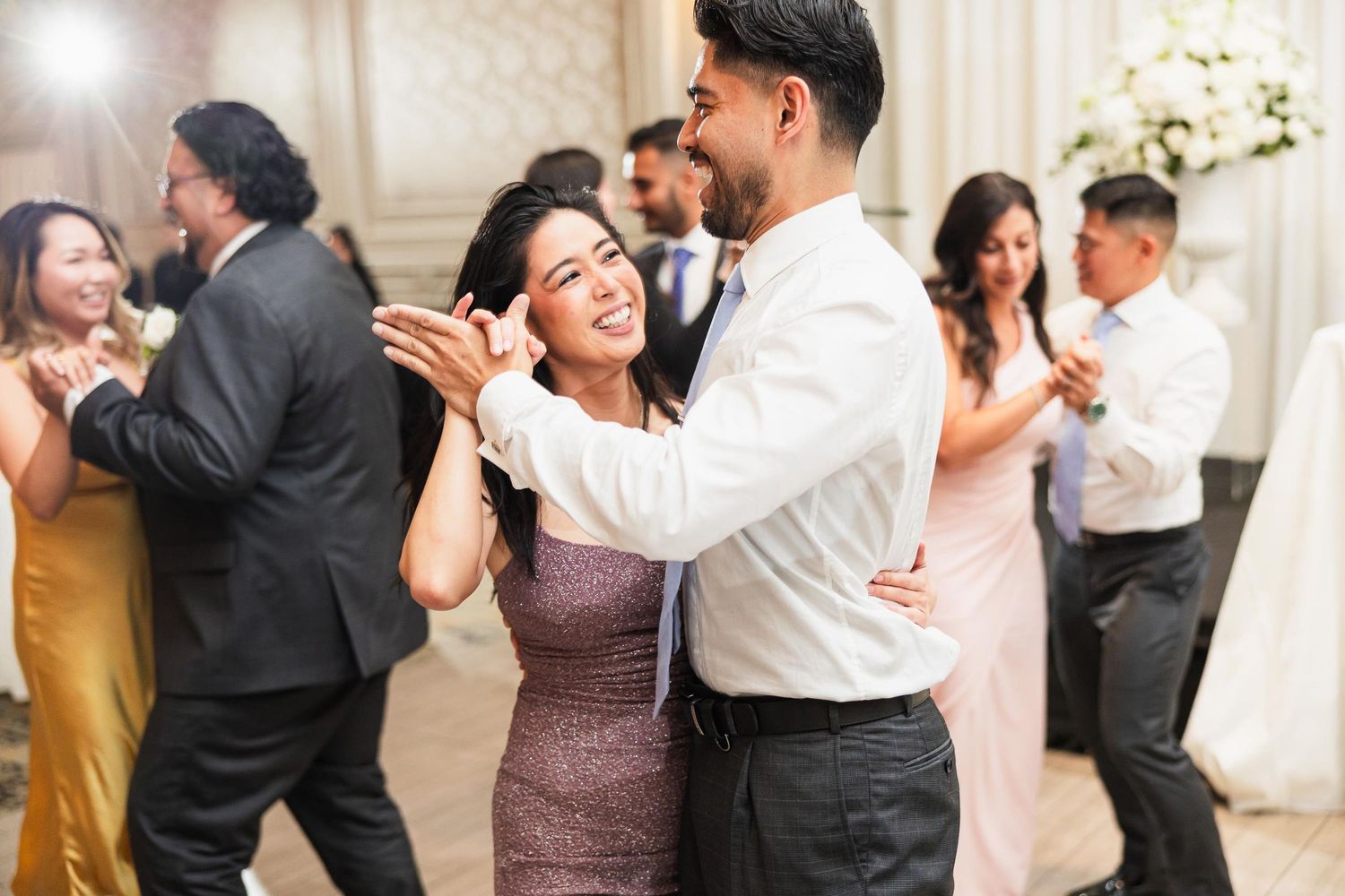 Wedding guests dance and laugh together on the dance floor at an elegant reception with white floral decorations.
