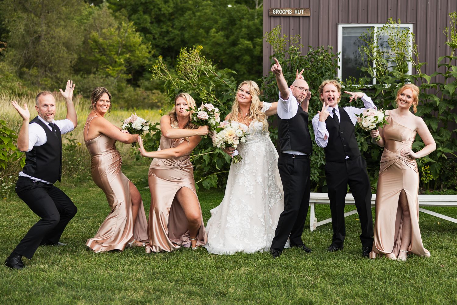 Wedding party having fun posing on grass in front of rustic barn with bridesmaids in blush dresses.