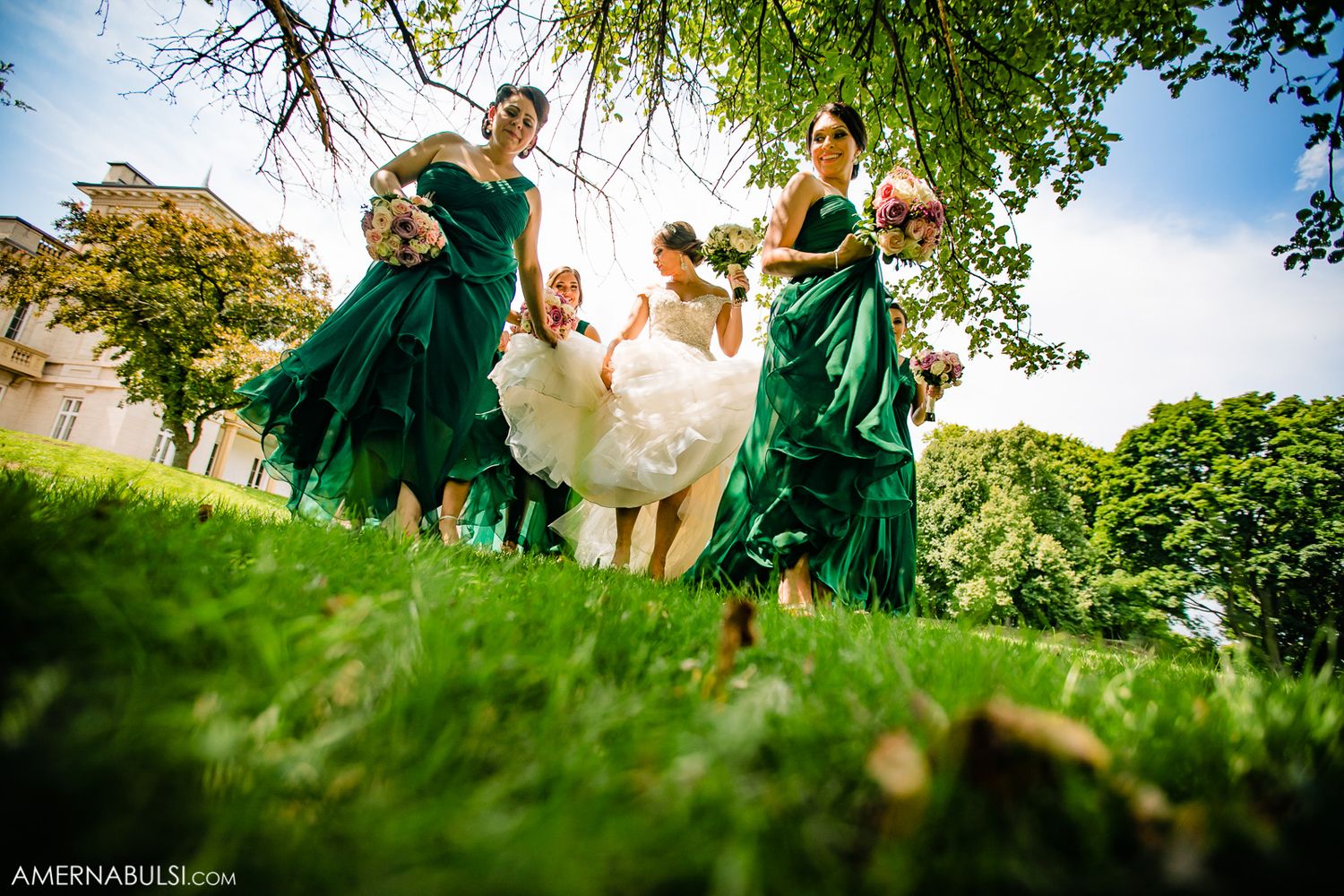 Wedding party in emerald green dresses walking through lush grass on a sunny day.