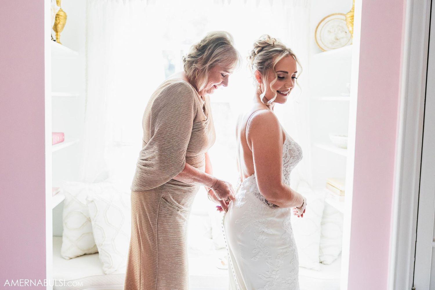 Two people getting ready, one helping adjust the back of a white wedding dress.
