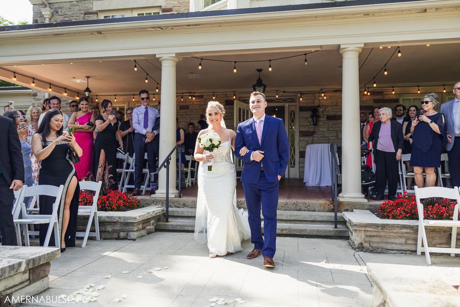 A formal wedding ceremony outdoors at a stone venue with white chairs and columns.
