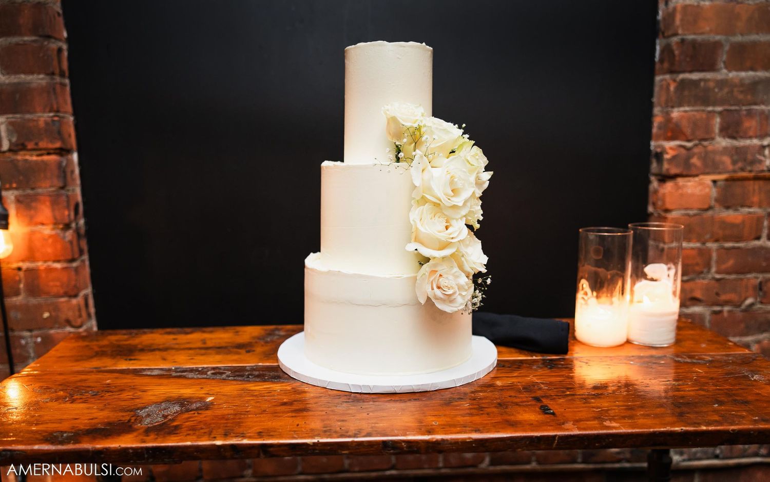 Elegant three-tier white wedding cake with fresh flowers on wooden table with candlelight.