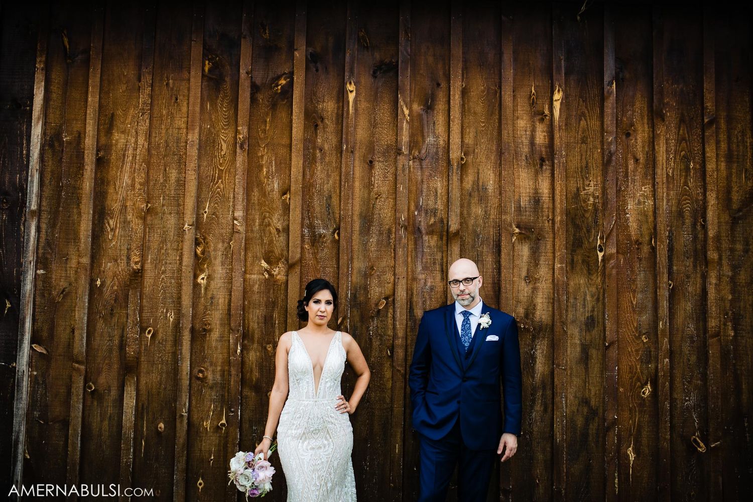 A wedding couple poses against a rustic wooden barn wall with a bridal bouquet.