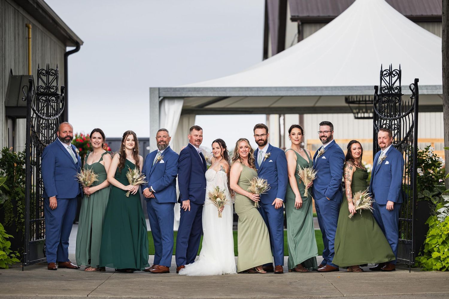 Wedding party in blue suits and green dresses poses formally against modern building.