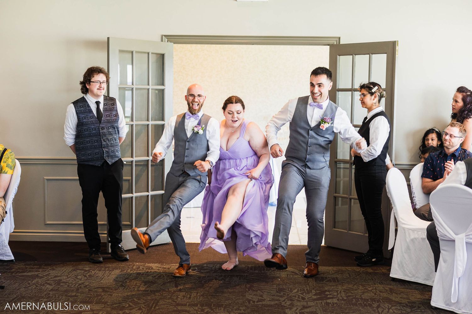 Wedding party members in purple dresses and gray vests pose playfully in indoor venue.