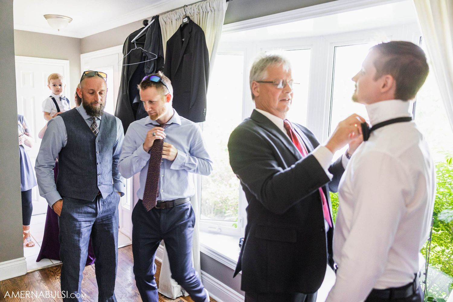 Men getting ready in formal attire before wedding ceremony in bright room with hanging suits.