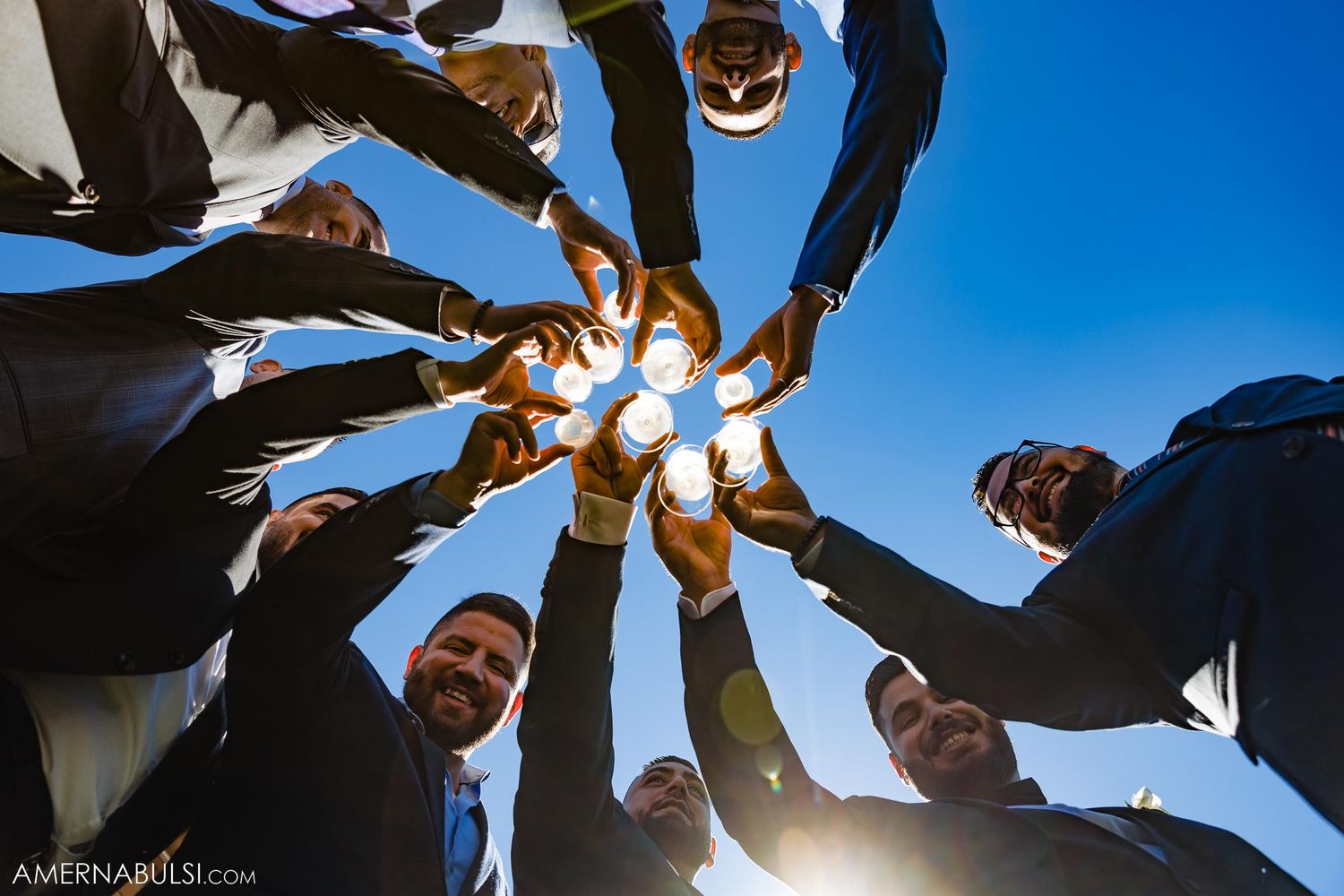Groomsmen create circle formation while toasting drinks against blue sky.