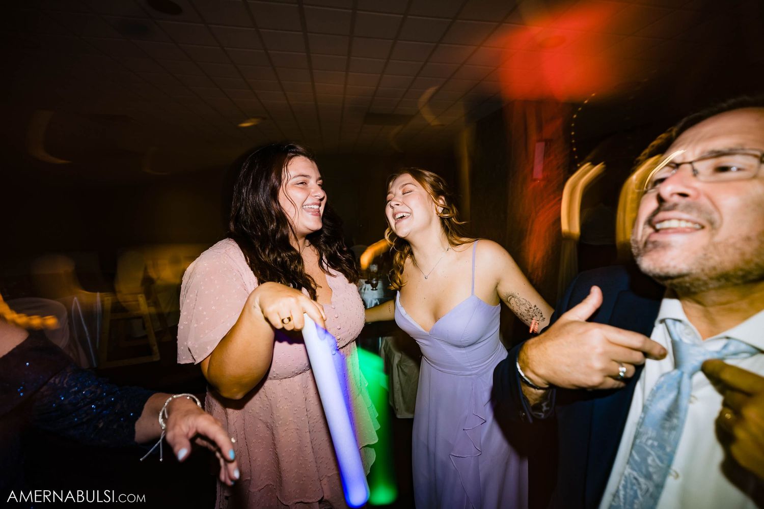 Wedding guests laugh and dance under colorful lights at reception party.