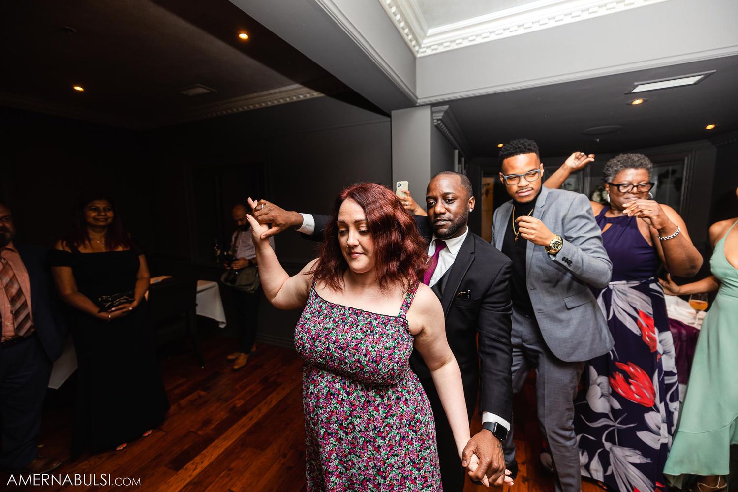 Wedding guests dancing in line on wooden dance floor at indoor reception.