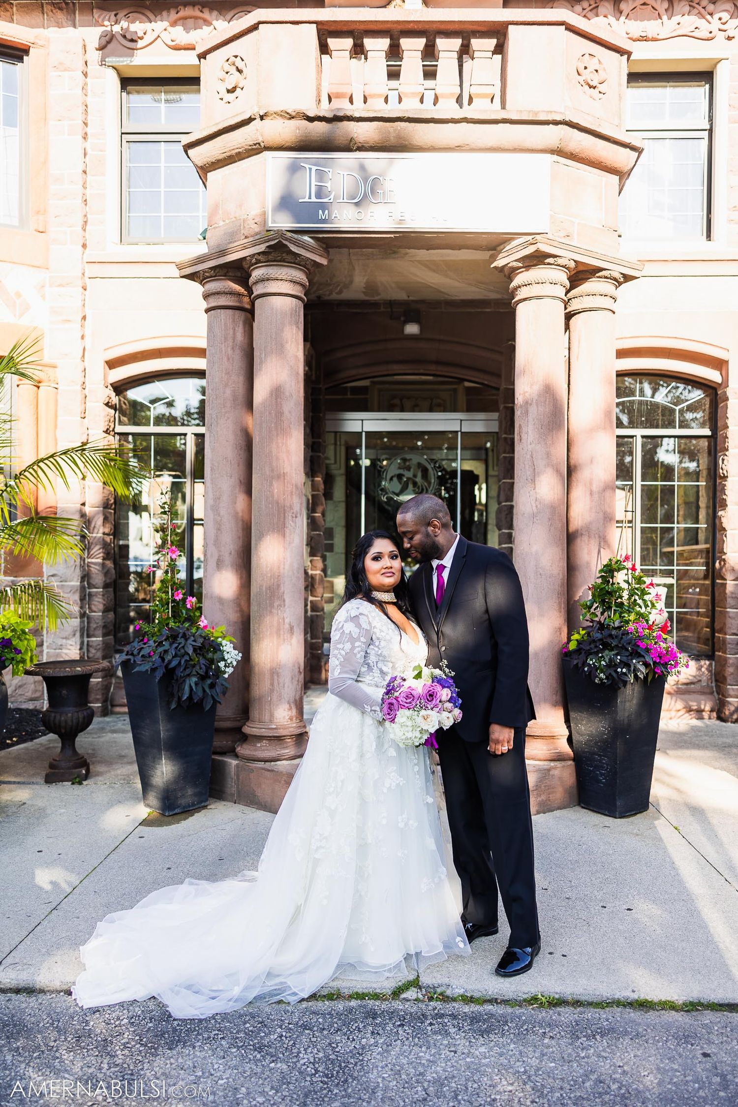 A couple poses in wedding attire under an ornate stone entrance with decorative pillars and flower planters.