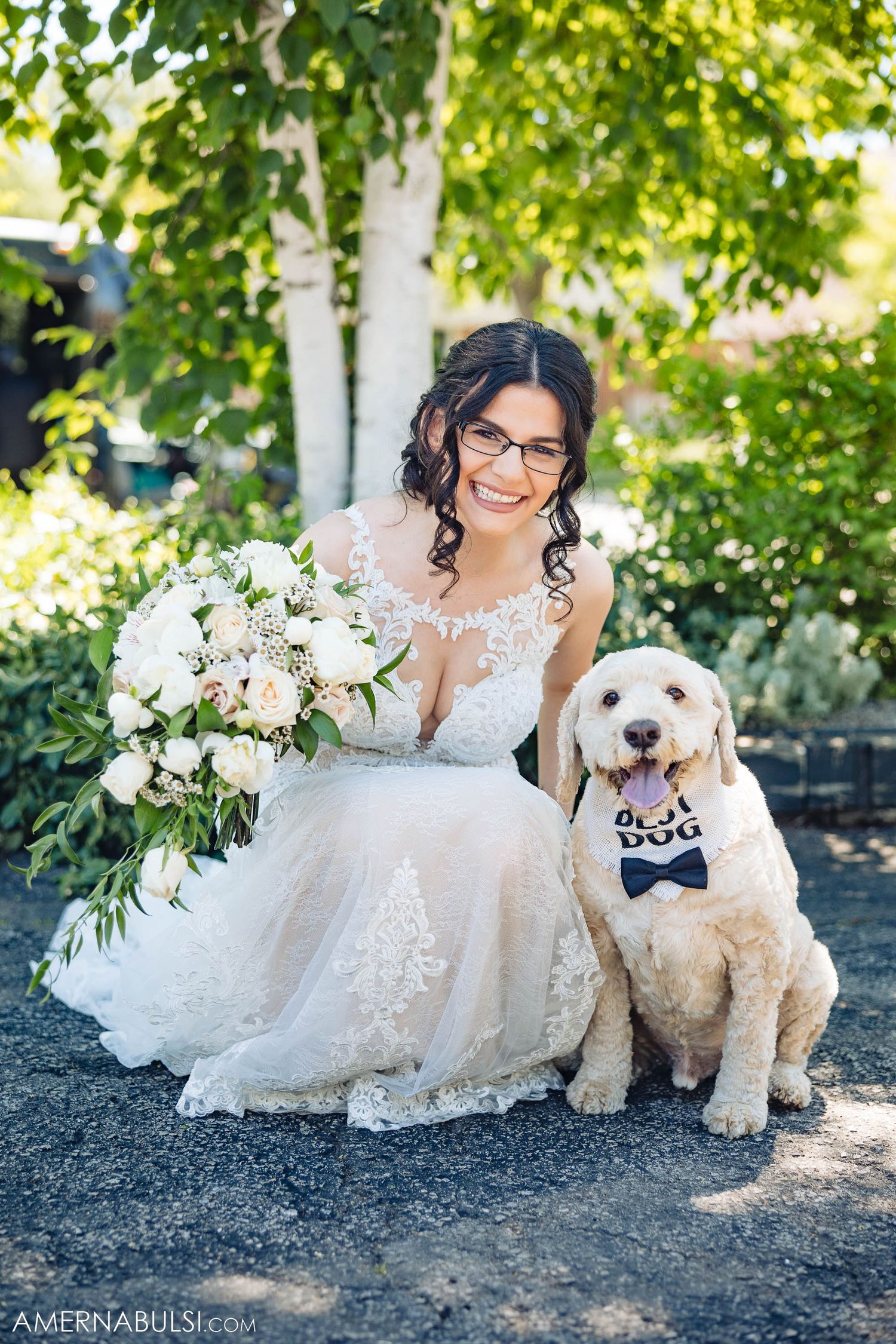 A bride in a lace wedding dress poses with her goldendoodle dog wearing a bow tie in a garden setting.