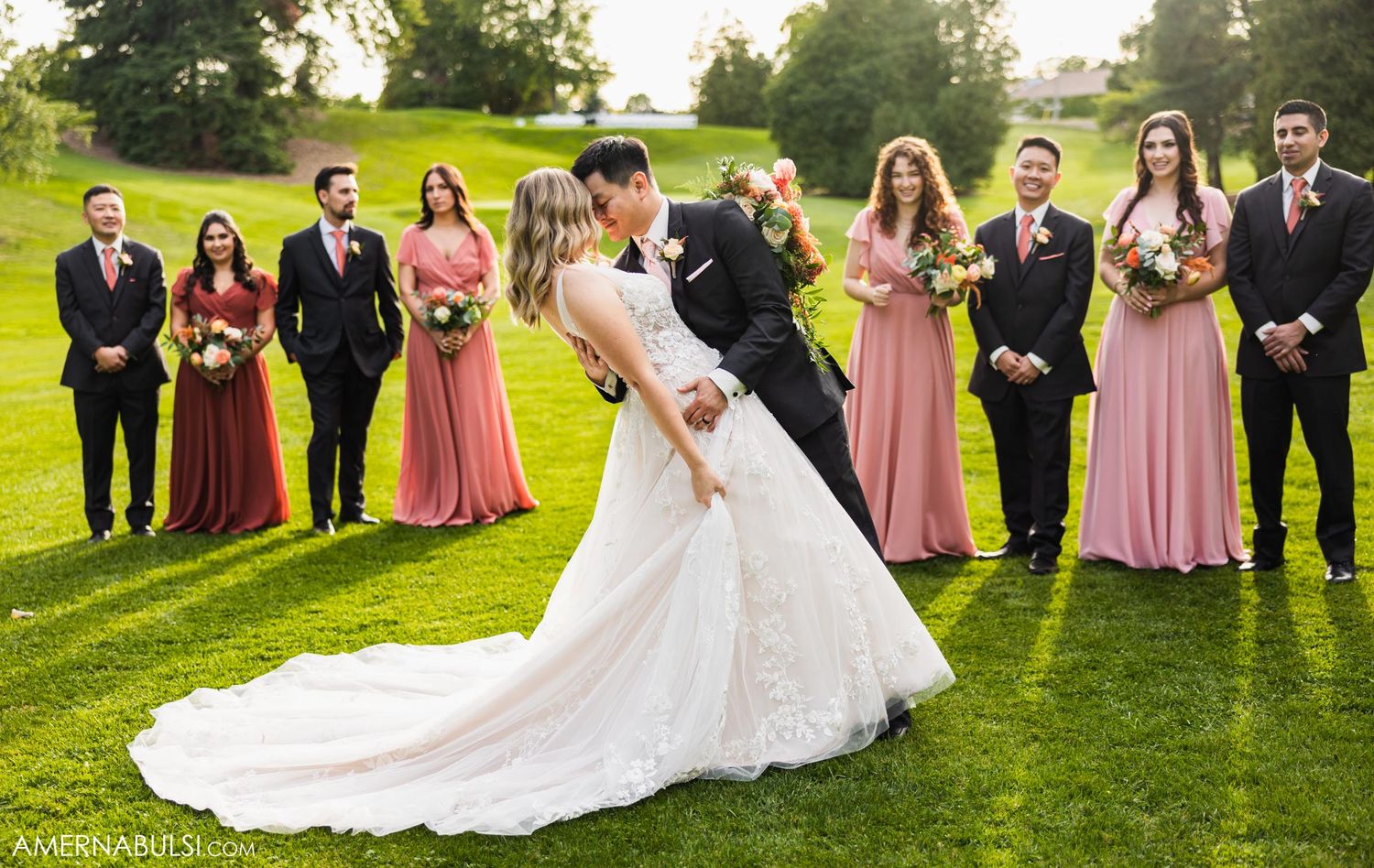 Wedding party stands in formation on lush green lawn while couple shares romantic dip pose.