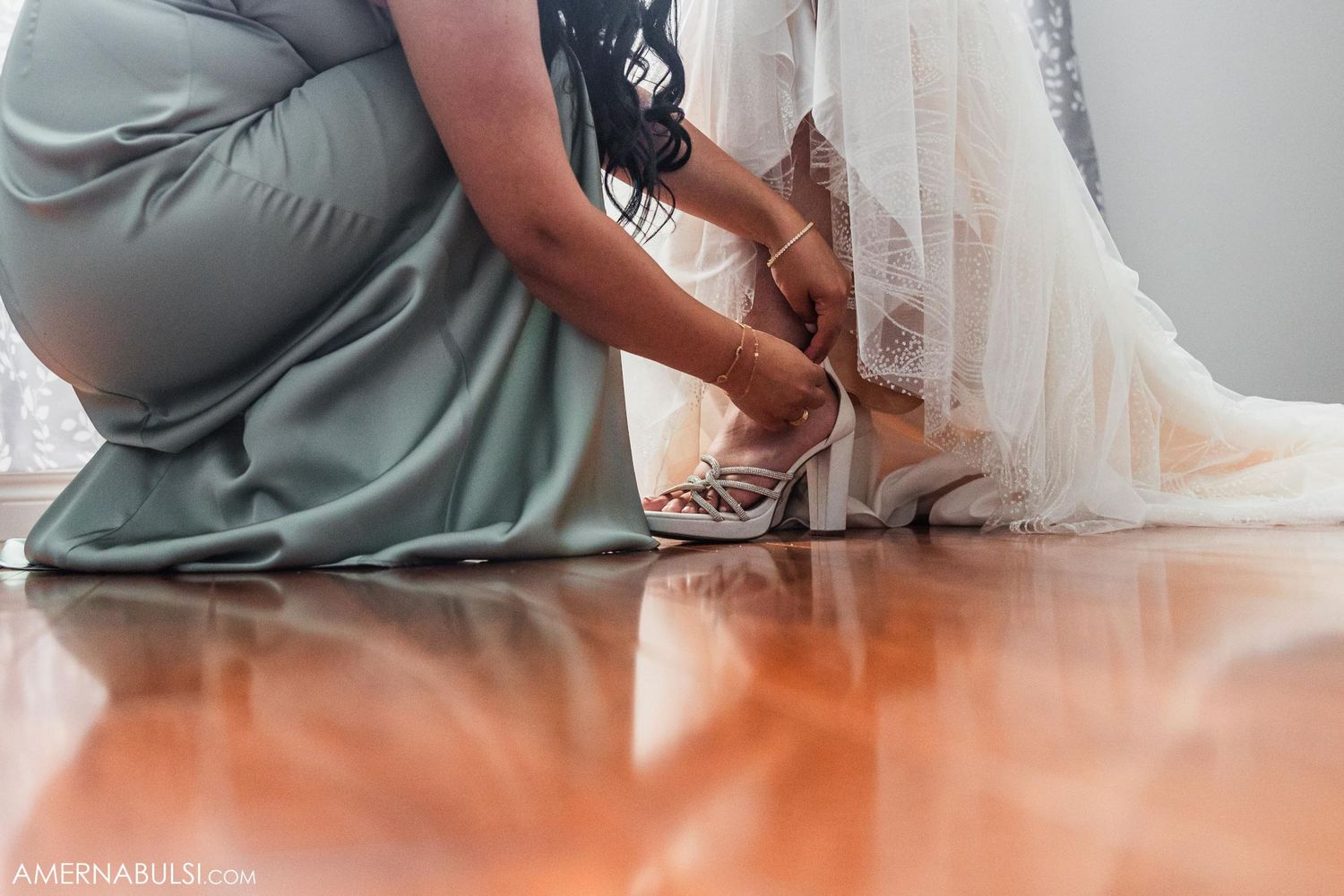 Low angle view of sage green dress and white wedding gown hem on hardwood floor.