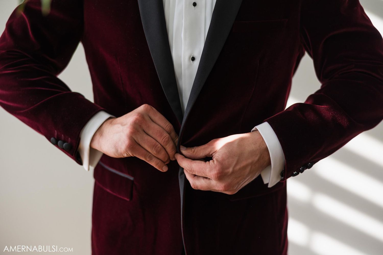Close-up of hands buttoning a burgundy velvet tuxedo jacket with black lapels.