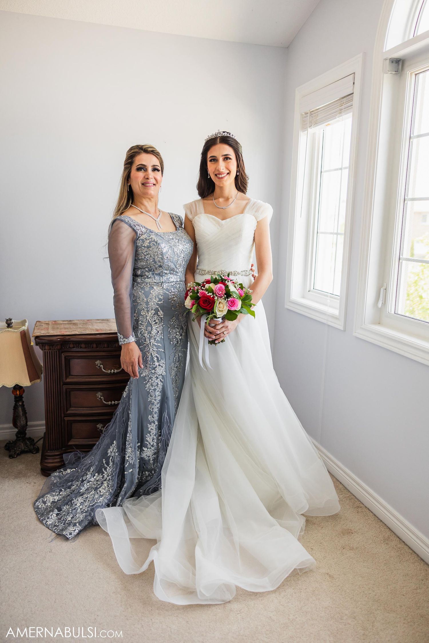 Two women in formal gowns pose together in an elegant room with natural light.