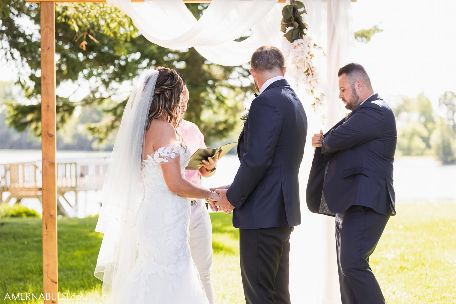 A wedding ceremony takes place under a wooden pergola with white draping in an outdoor garden setting.