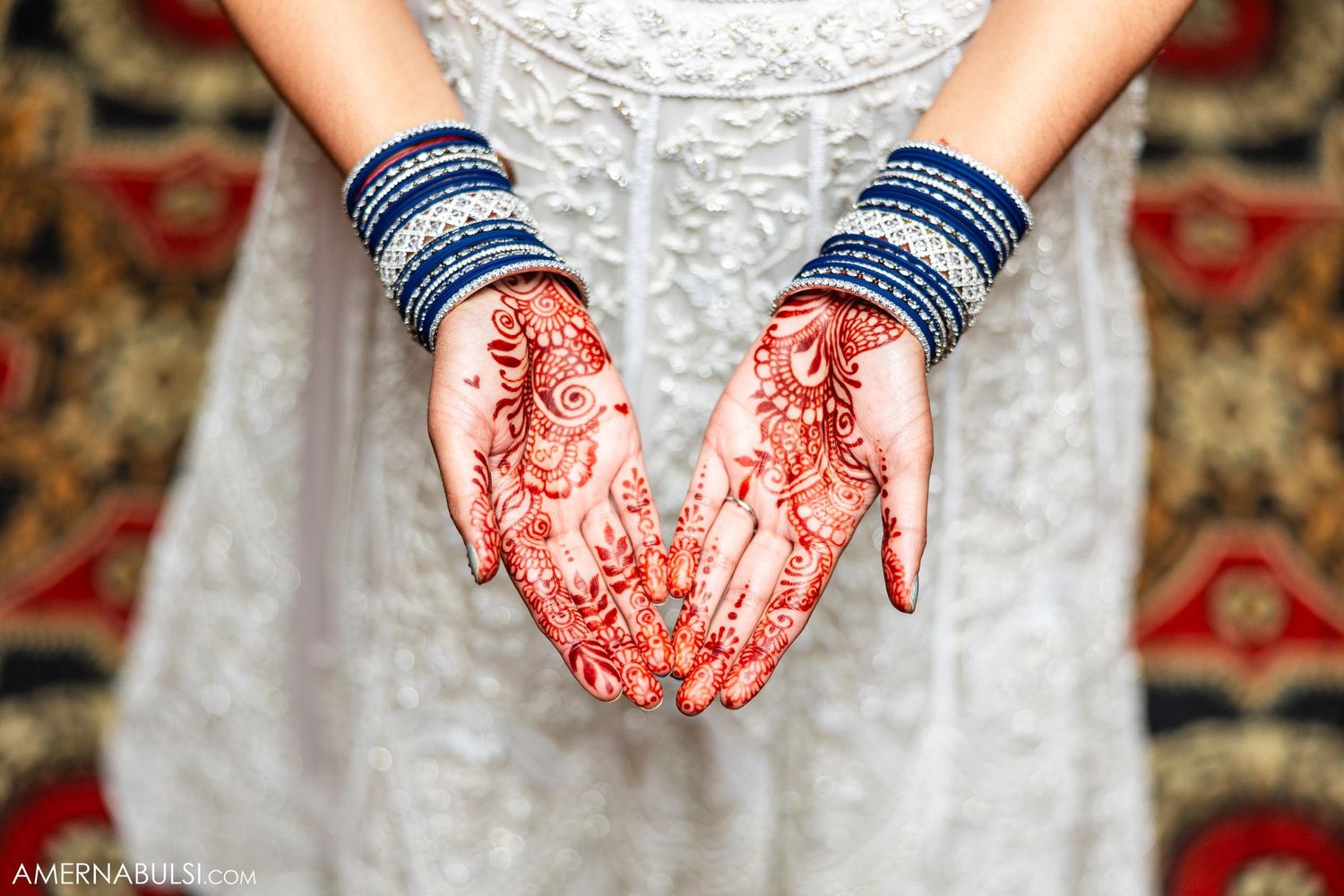 Close-up of intricate henna designs on hands with blue bangles against white fabric.