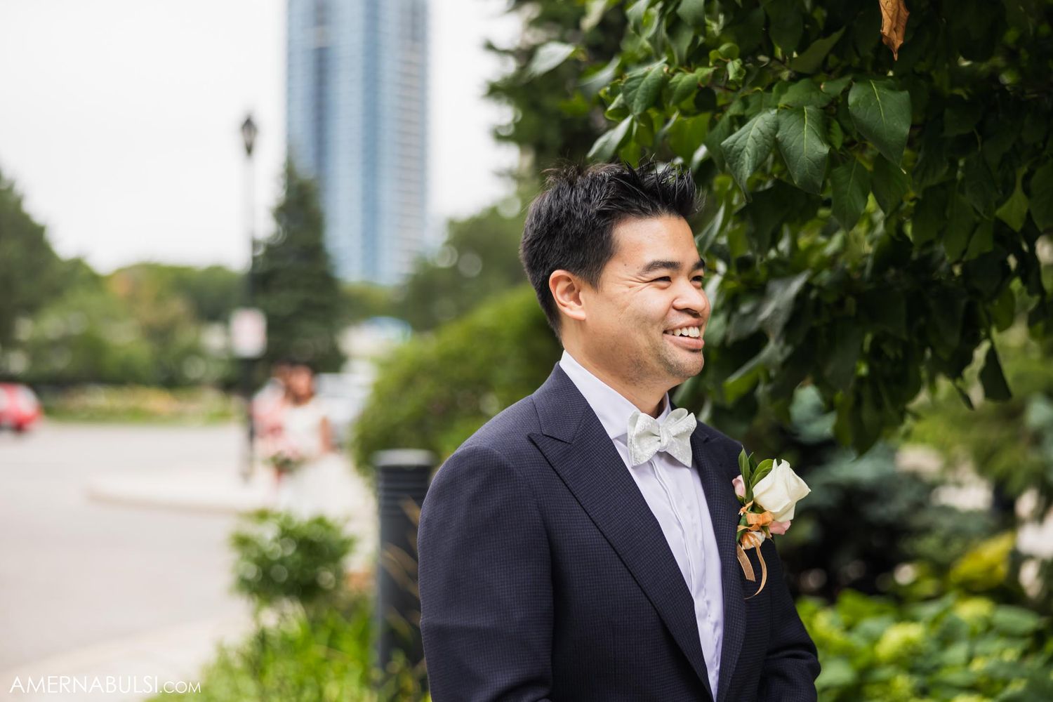 Groom in formal attire smiles outdoors with city buildings in background.