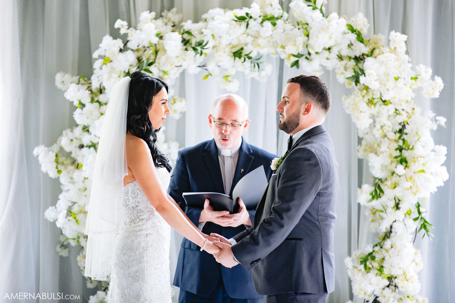 Wedding ceremony with white floral arch and couple exchanging vows in front of officiant.
