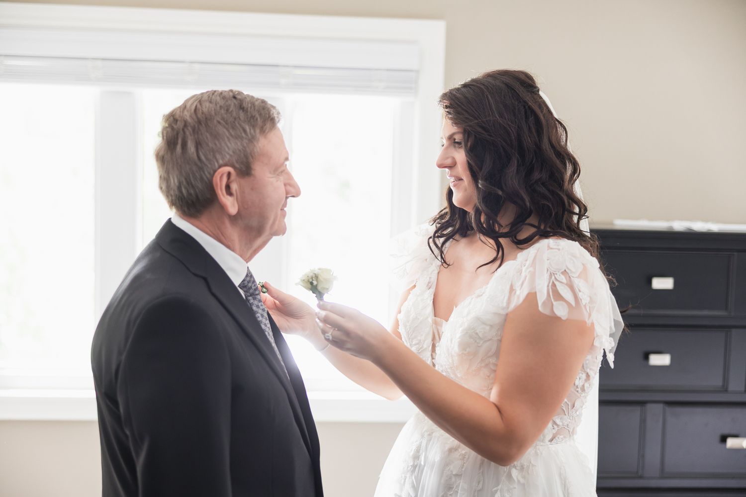 Bride in lace gown adjusting boutonniere on man in black suit before wedding ceremony.